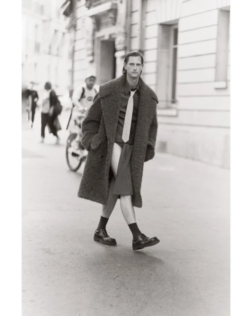 Monochrome street-style fashion shot: A male model in an oversized coat, shirt, tie, and shorts walks on a city street with a cyclist.