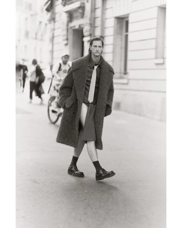 Monochrome street-style fashion shot: A male model in an oversized coat, shirt, tie, and shorts walks on a city street with a cyclist.