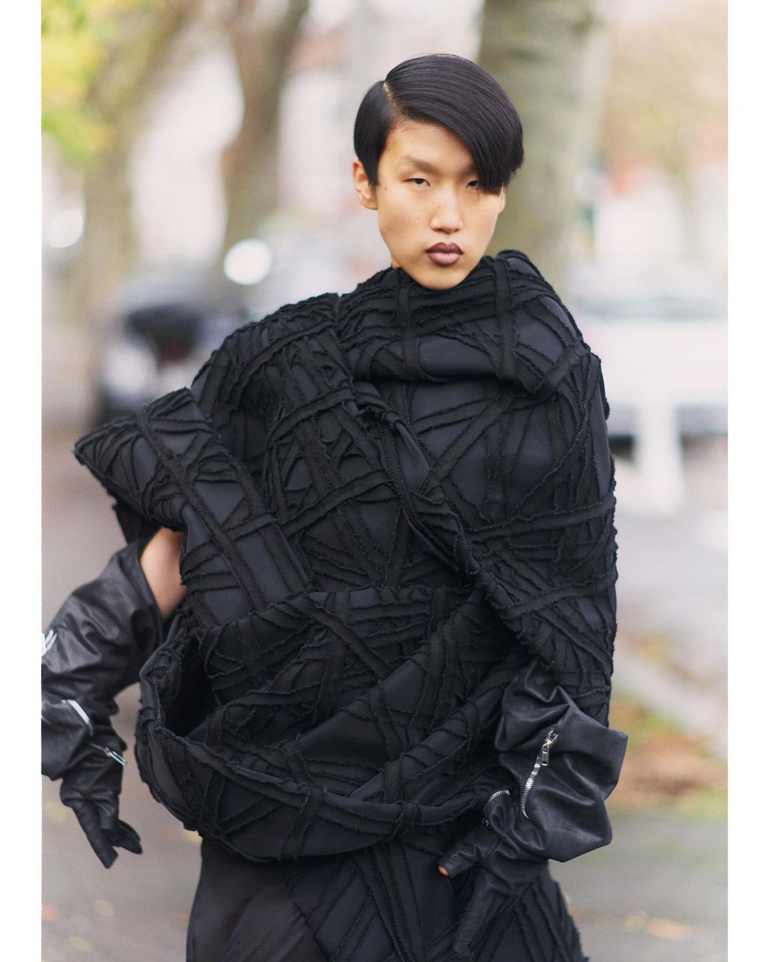 Person wearing a unique black woven garment standing outdoors with cars and foliage blurred in the background. Unusual fashion statement.