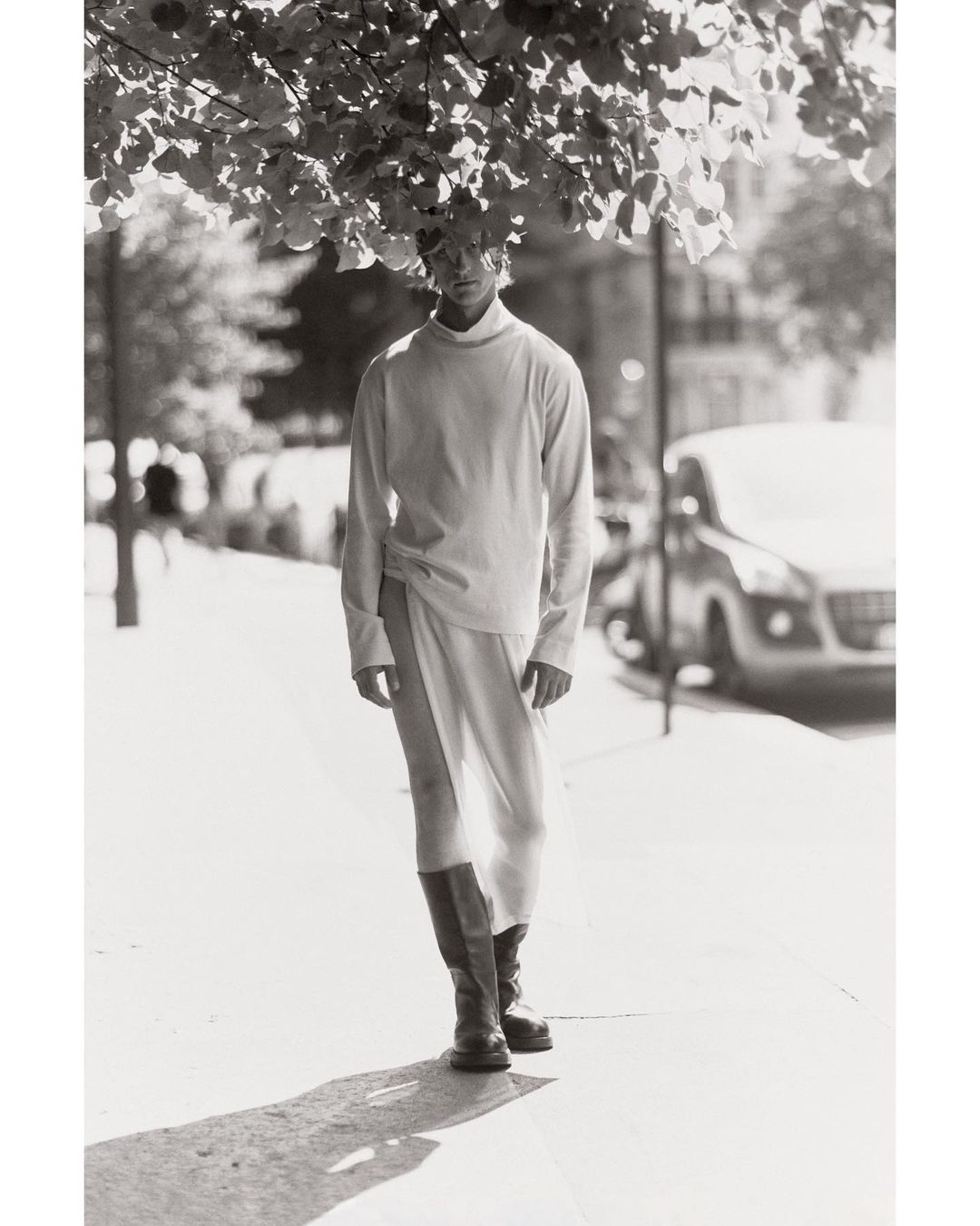 Male fashion model posing outdoors on a city street wearing long-sleeved shirt, skirt with slit, and boots under tree canopy.