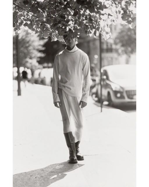 Male fashion model posing outdoors on a city street wearing long-sleeved shirt, skirt with slit, and boots under tree canopy.