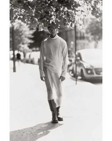 Male fashion model posing outdoors on a city street wearing long-sleeved shirt, skirt with slit, and boots under tree canopy.