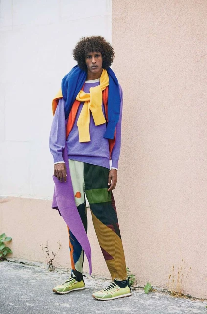 A stylish adult male model is posing outdoors wearing a purple sweater, colorful geometric patterned pants, and chartreuse-colored sneakers.