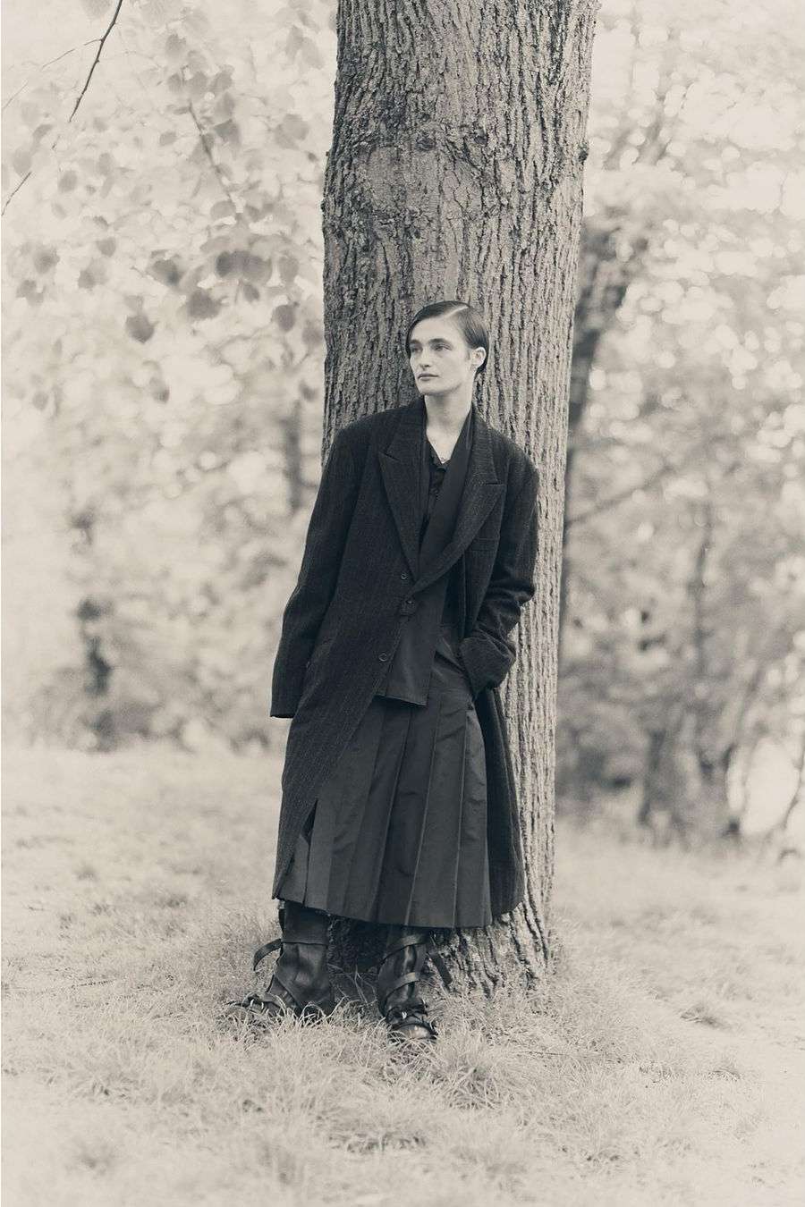 Monochrome portrait of a person in a coat and kilt, leaning against a tree in a grassy outdoor setting with blurred foliage.