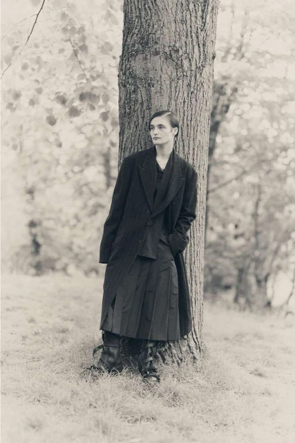 Monochrome portrait of a person in a coat and kilt, leaning against a tree in a grassy outdoor setting with blurred foliage.