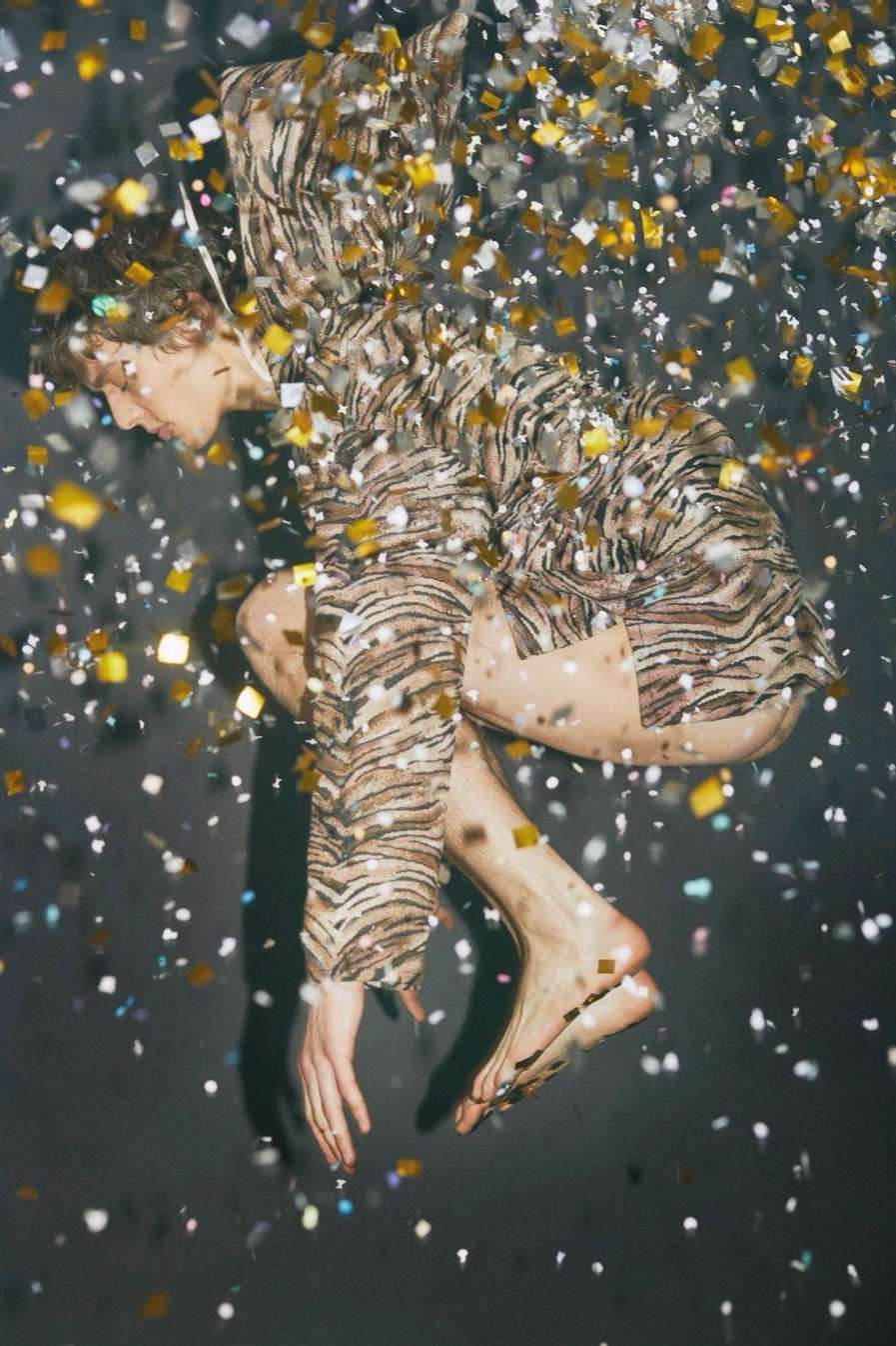 Barefoot young adult in tiger-striped shirt and shorts reclines amid shower of golden confetti against dark background in studio setting.