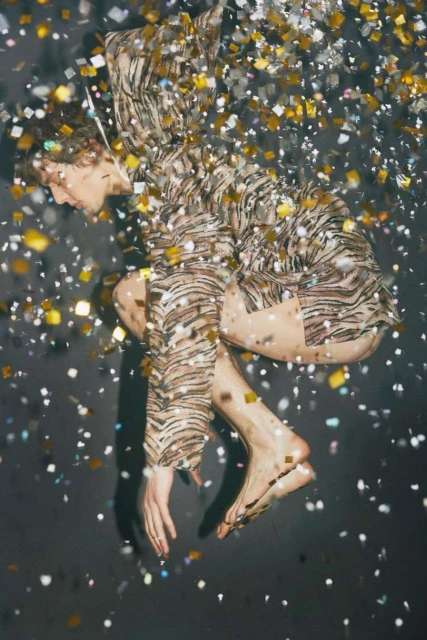 Barefoot young adult in tiger-striped shirt and shorts reclines amid shower of golden confetti against dark background in studio setting.