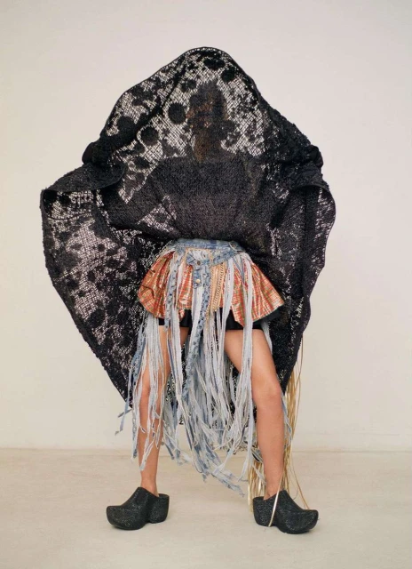 Person wearing an elaborate skirt and lace covering posing in front of a white wall inside a photo studio, studio lighting.