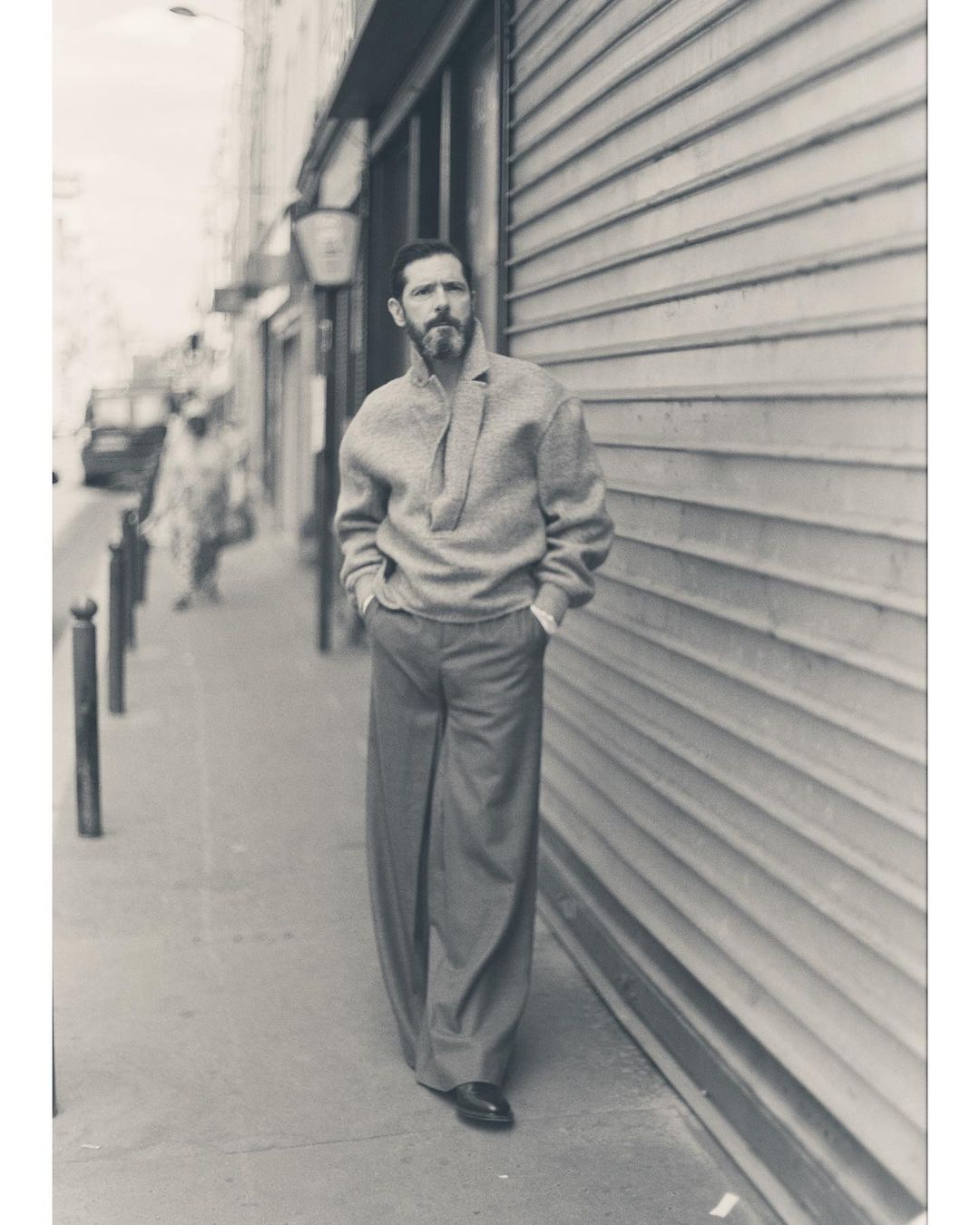 Man with beard wearing textured sweater and wide-leg trousers walking on a Paris sidewalk next to a metal shutter