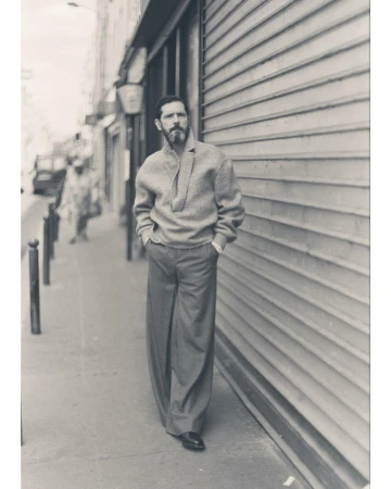 Man with beard wearing textured sweater and wide-leg trousers walking on a Paris sidewalk next to a metal shutter