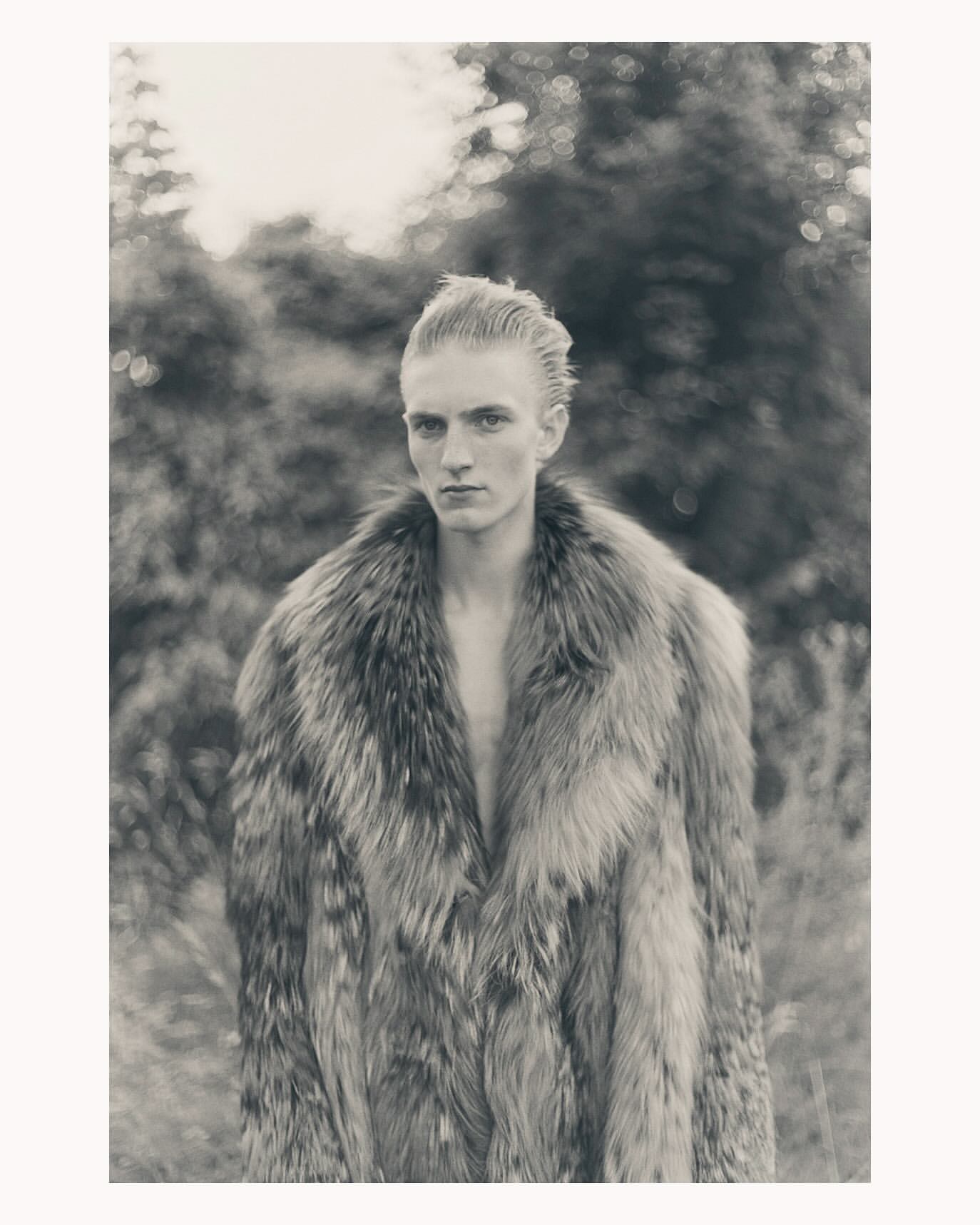 Monochrome portrait of a fair-skinned adult male in a luxurious fur coat, posing outdoors with a blurred forest background, creating a vintage feel.