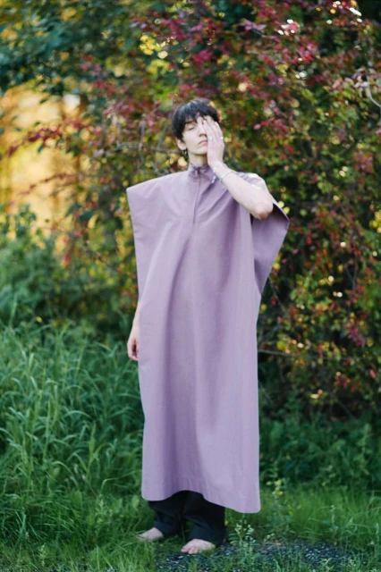 A young adult in a lavender dress stands barefoot in a field of green grass near leafy bushes and trees in a natural outdoor setting.