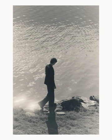 A man in a dark suit walking along a rocky shoreline beside water, photographed by Ola Rindal in Paris.