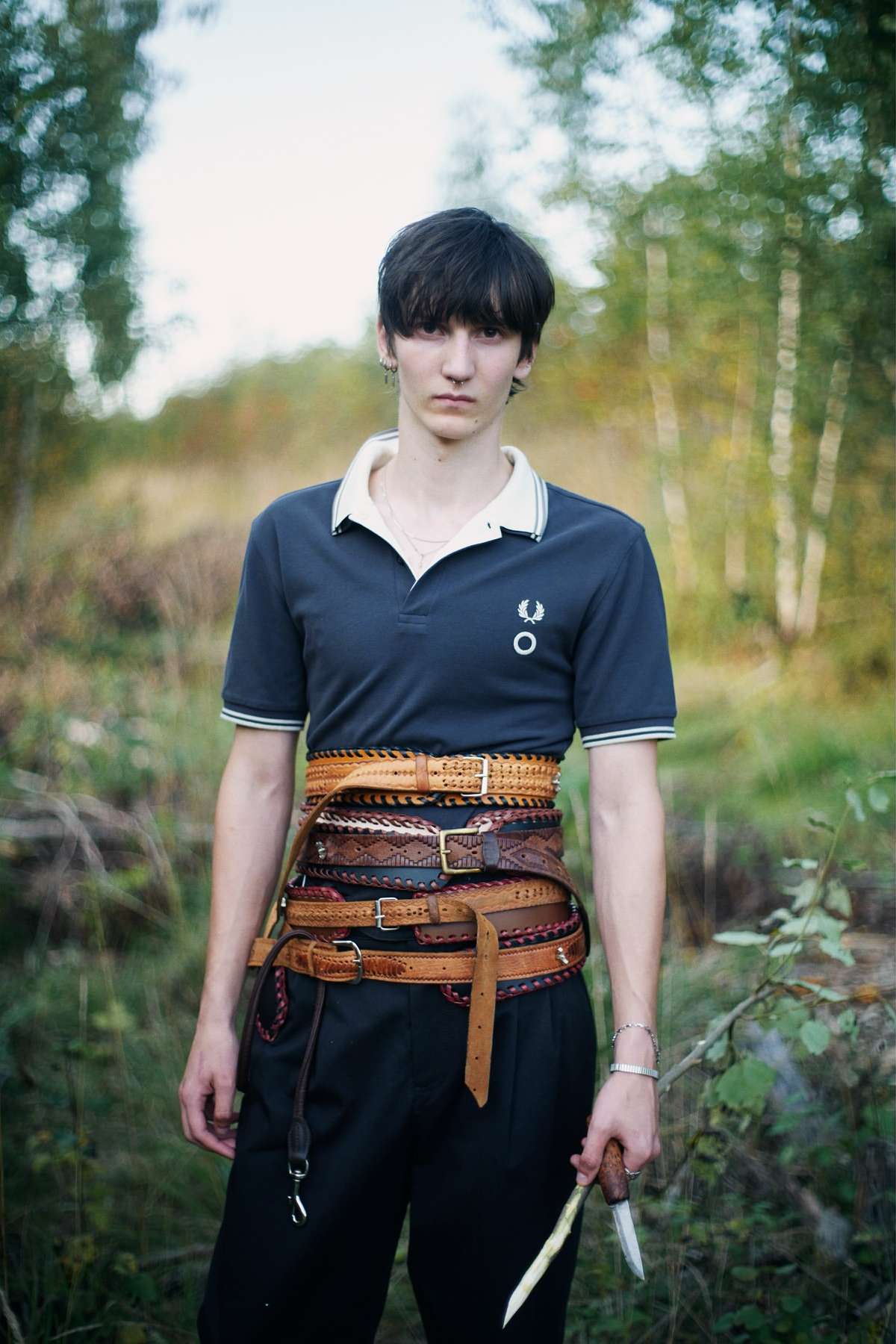 A person wearing a polo shirt and multiple belts stands in a wooded area holding a knife, looking directly at the camera in a medium shot.