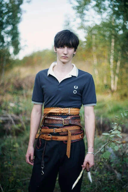 A person wearing a polo shirt and multiple belts stands in a wooded area holding a knife, looking directly at the camera in a medium shot.