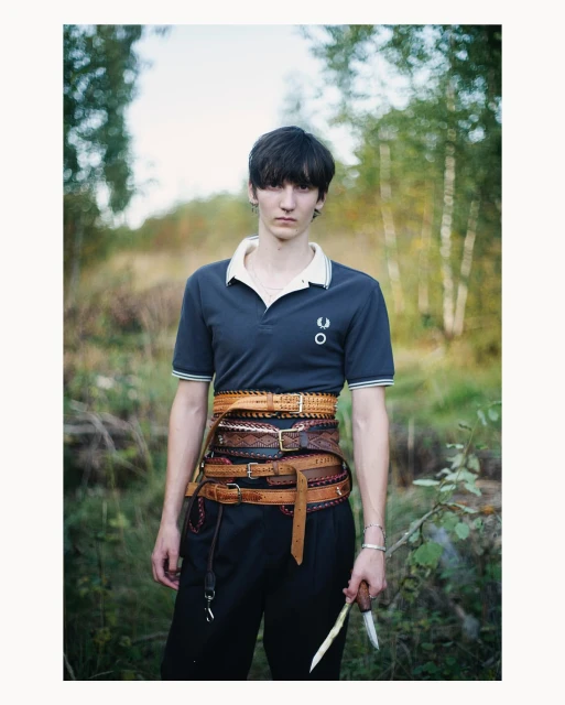 Portrait of a person wearing a blue polo shirt and several brown leather belts while standing in an outdoor grassy environment.