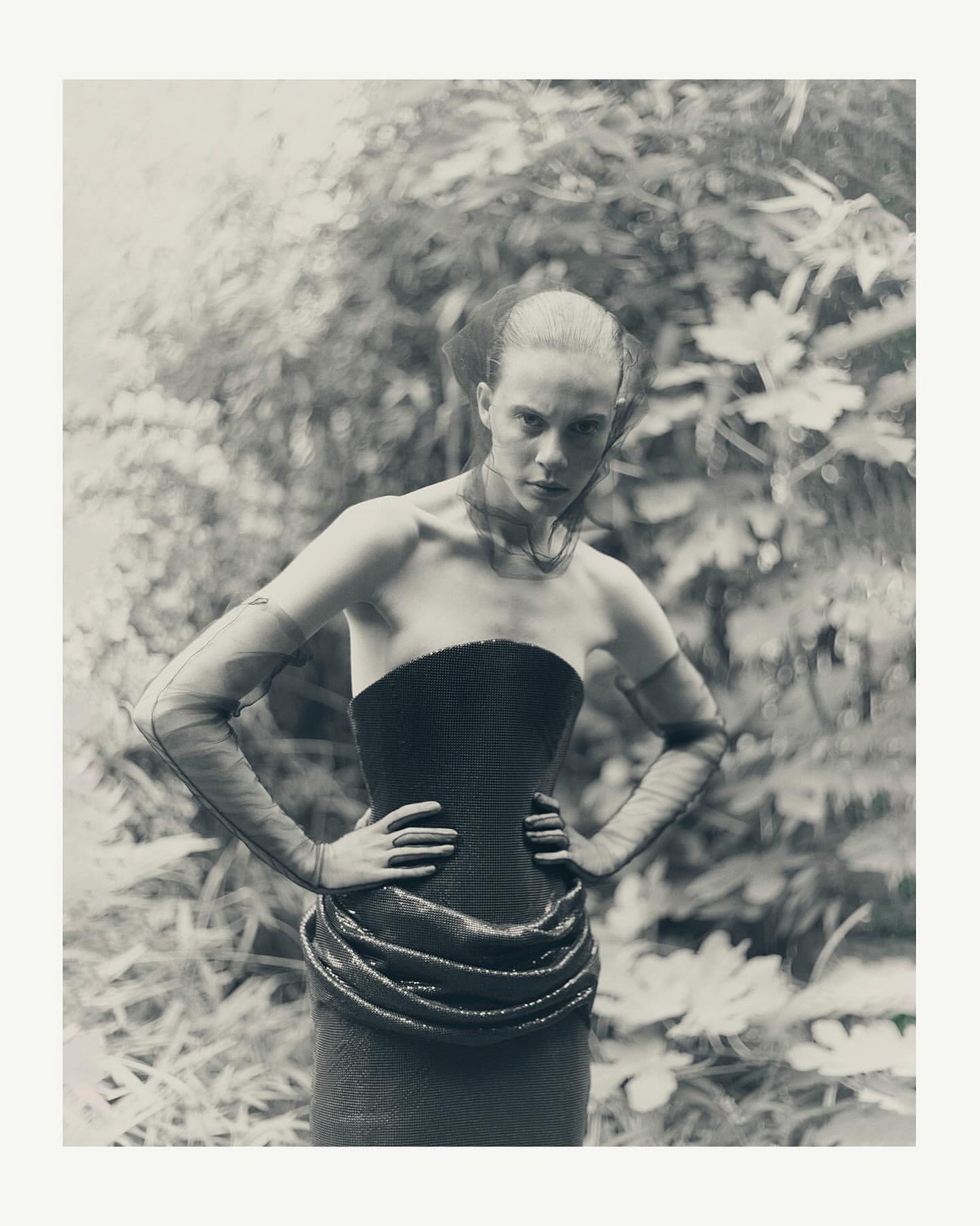 Monochrome image of a female fashion model in a strapless corset dress, tulle veil, and arm coverings, posed outdoors with foliage.