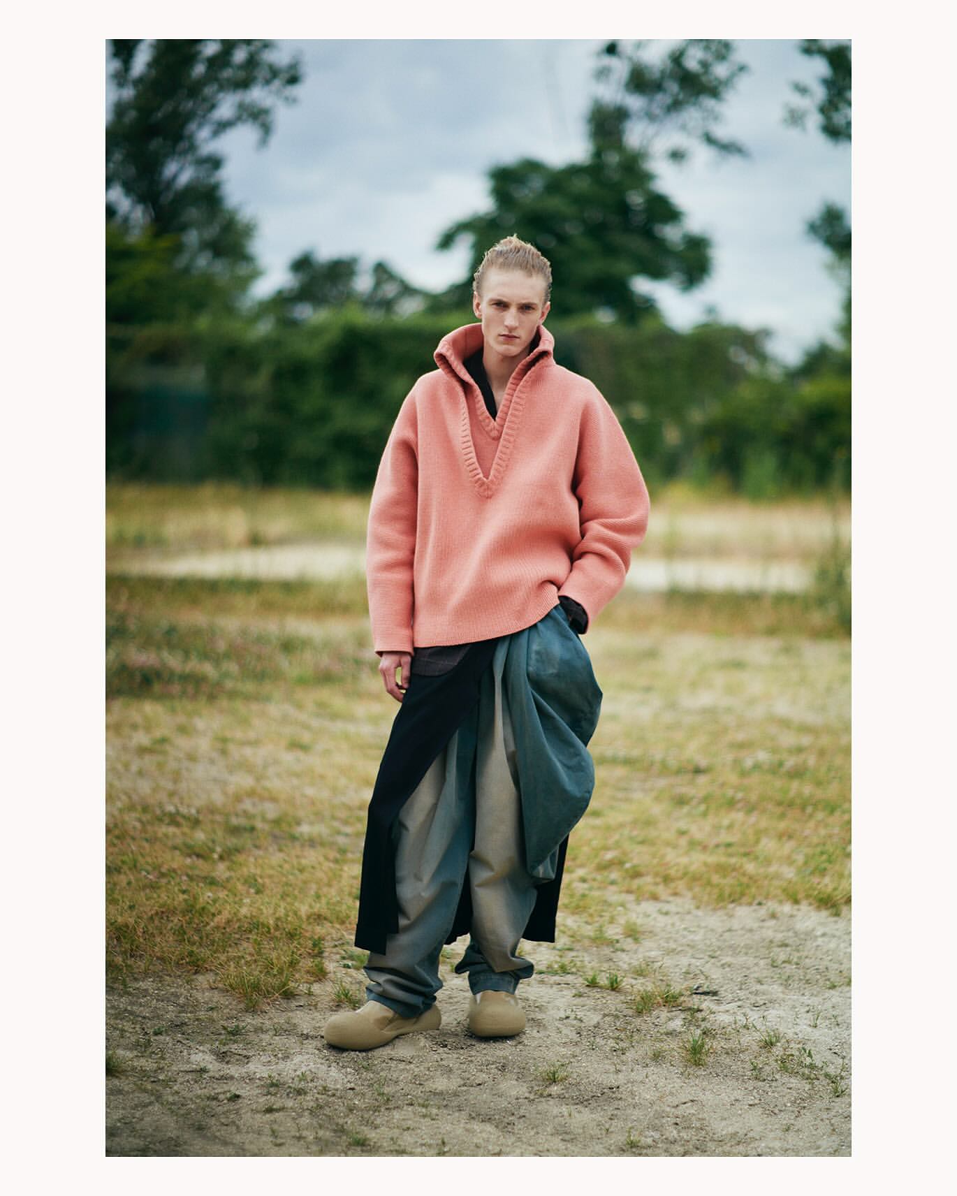 A person stands in a field with green and dry grasses, wearing a peach-colored sweater, blue pants, and tan boots. Overcast sky.