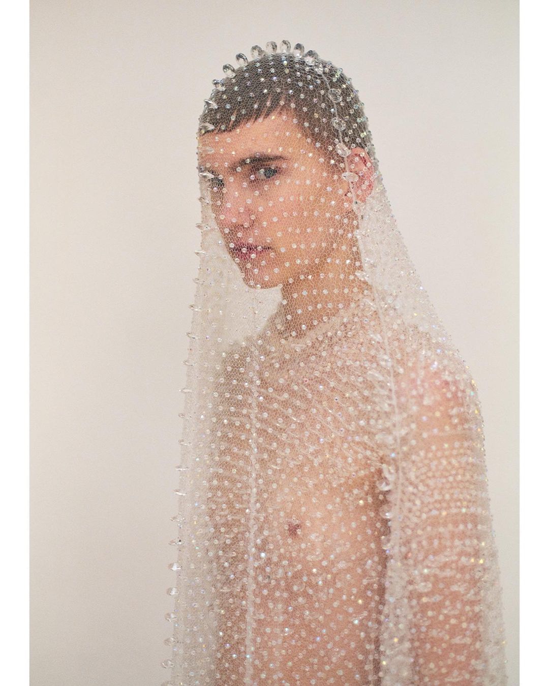Young adult poses in a beaded net top and headpiece against a plain background in a beauty and fashion photography studio setting.