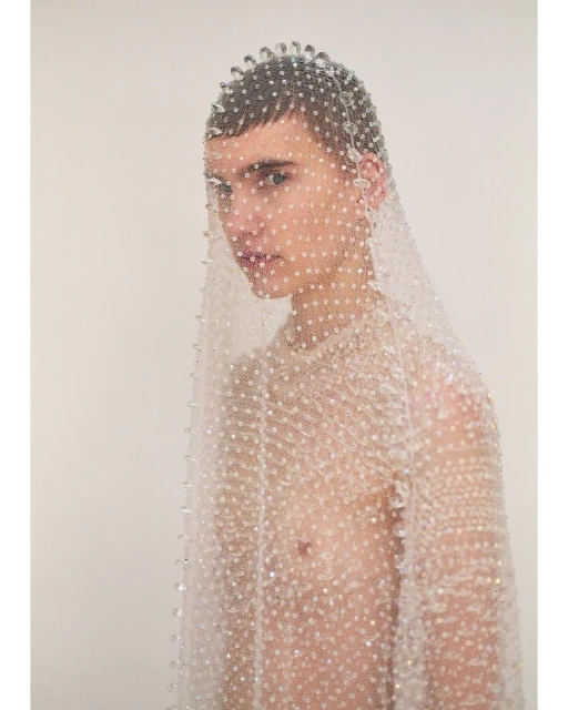 Young adult poses in a beaded net top and headpiece against a plain background in a beauty and fashion photography studio setting.