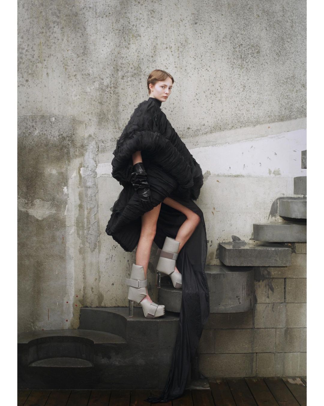 Fashion shot of a woman in a voluminous black dress with gray platform boots on stone steps in front of a weathered wall.