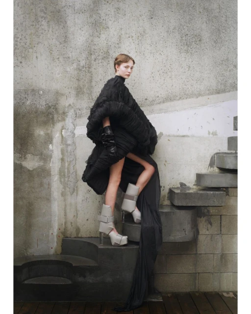 Fashion shot of a woman in a voluminous black dress with gray platform boots on stone steps in front of a weathered wall.