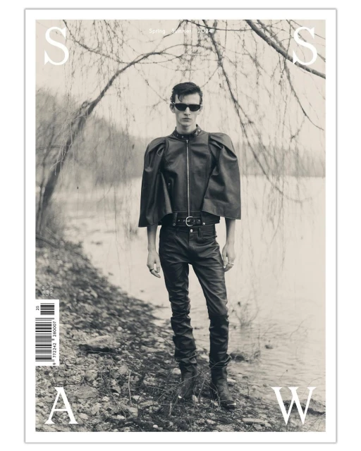 Monochrome magazine cover featuring a male model in leather clothing standing by a body of water with trees, promoting a spring/summer issue.