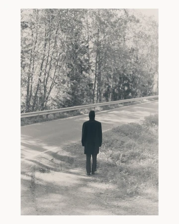 Monochromatic view of an adult male walking along a roadside near a forest with a dark coat and trousers, captured in a full shot.