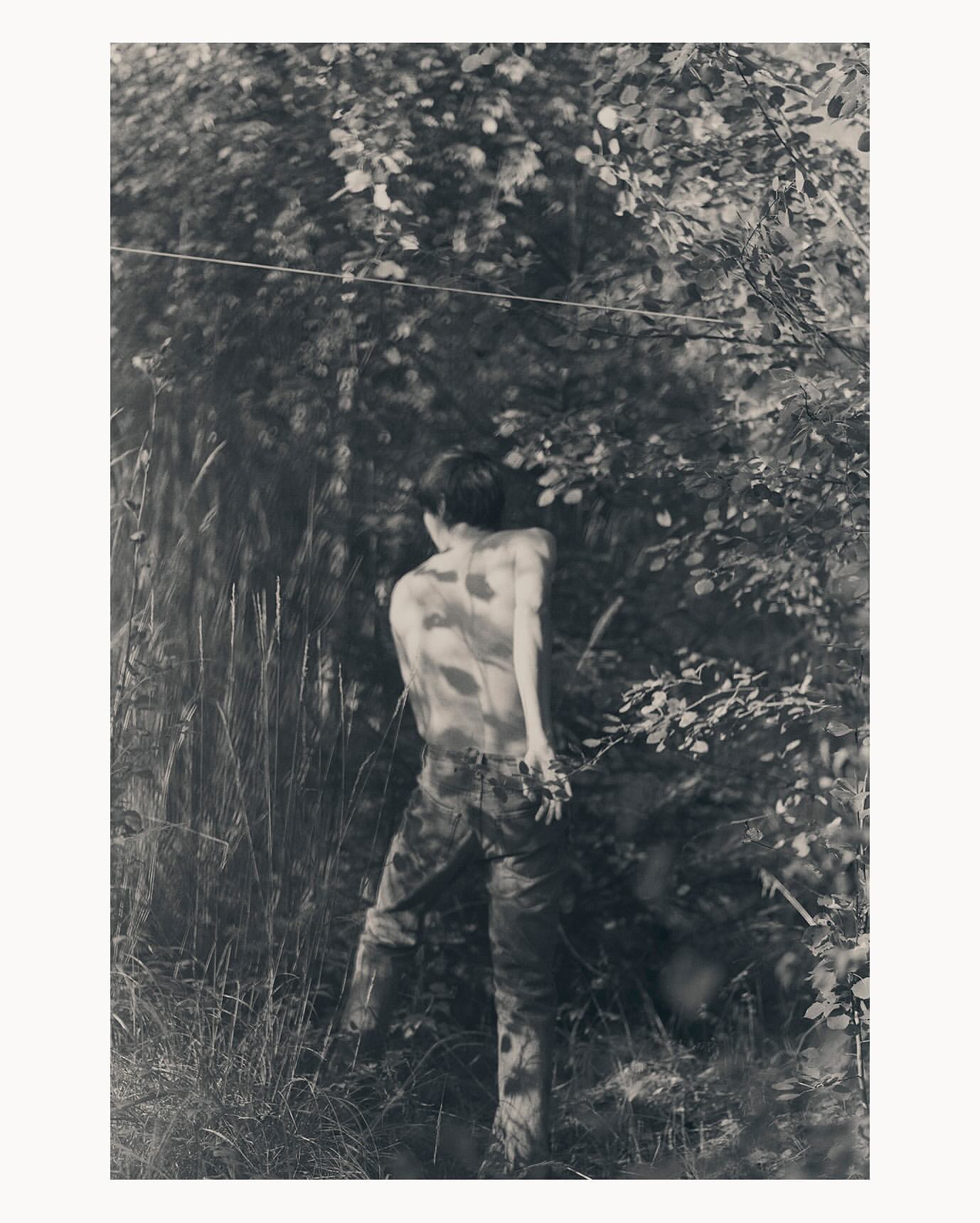 Monochrome photograph of a young adult outdoors with camouflage pants and no shirt, surrounded by grass, leaves, and trees.