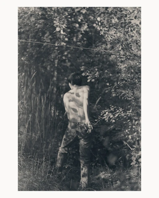 Monochrome photograph of a young adult outdoors with camouflage pants and no shirt, surrounded by grass, leaves, and trees.