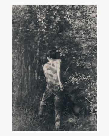 Monochrome photograph of a young adult outdoors with camouflage pants and no shirt, surrounded by grass, leaves, and trees.