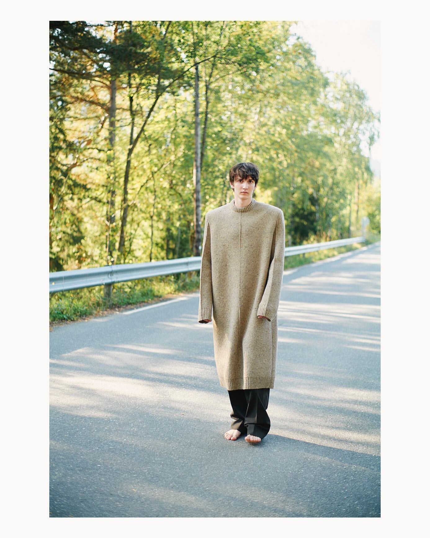 A young adult in an oversized sweater and dark pants stands barefoot in the middle of an asphalt road with a wooded background.