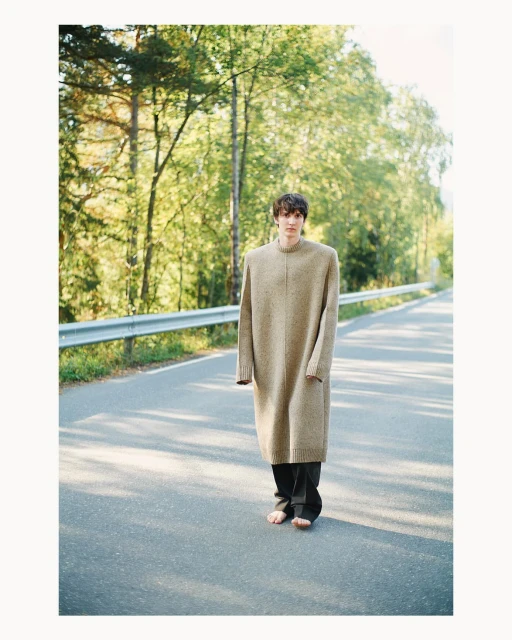 A young adult in an oversized sweater and dark pants stands barefoot in the middle of an asphalt road with a wooded background.