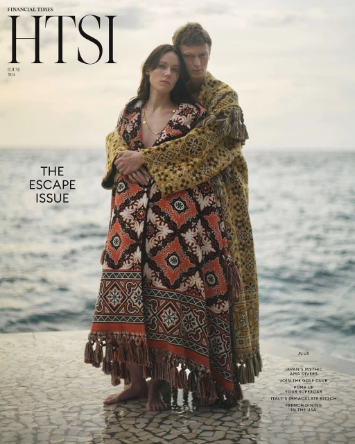 A couple stands on a stone platform with the ocean behind them. They are draped in ornate, patterned textiles in front of a Financial Times magazine cover.