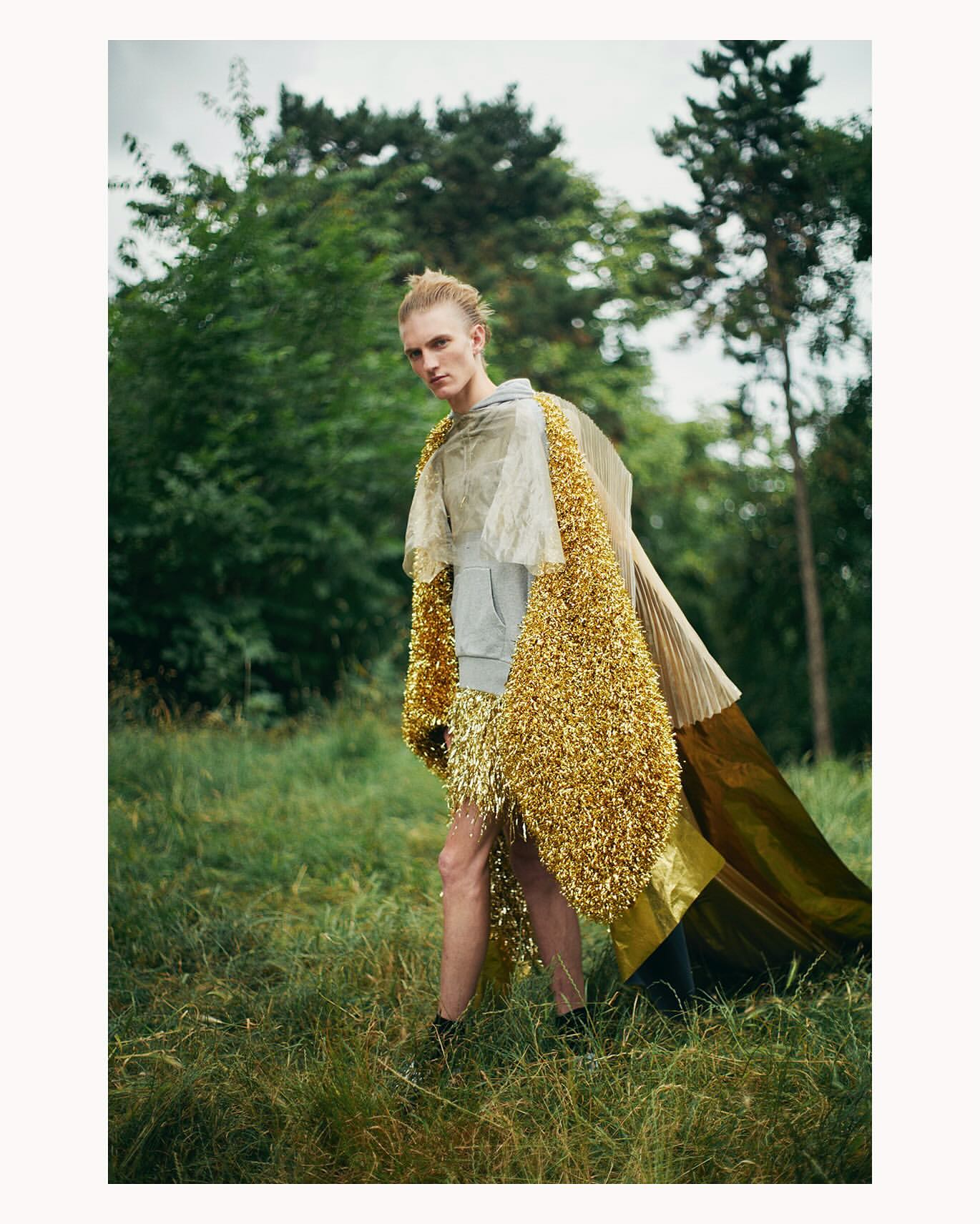 Person wearing a gold tinsel cloak stands in a grassy, outdoor area surrounded by trees. Fashion editorial, nature background.