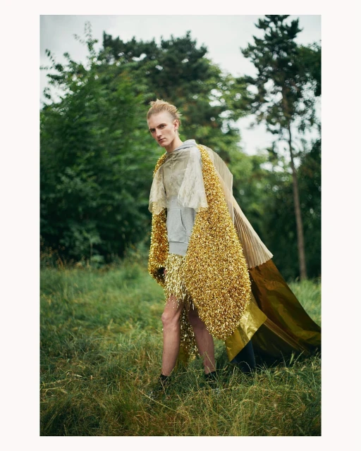 Person wearing a gold tinsel cloak stands in a grassy, outdoor area surrounded by trees. Fashion editorial, nature background.