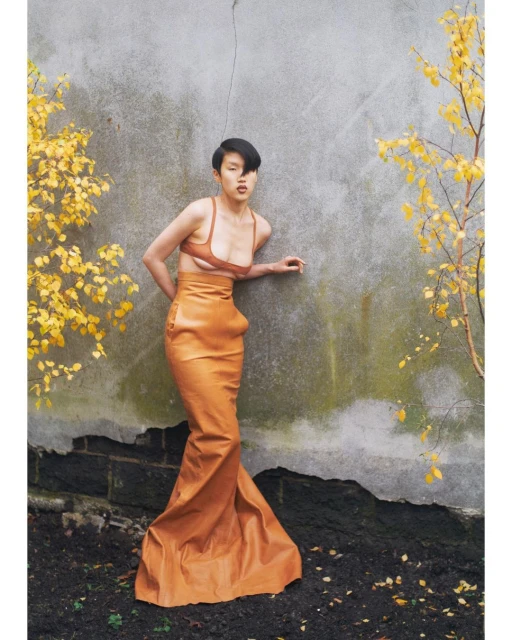 Woman in an orange dress poses against a weathered wall with sparse yellow foliage; outdoor fashion shot, medium shot, natural lighting.