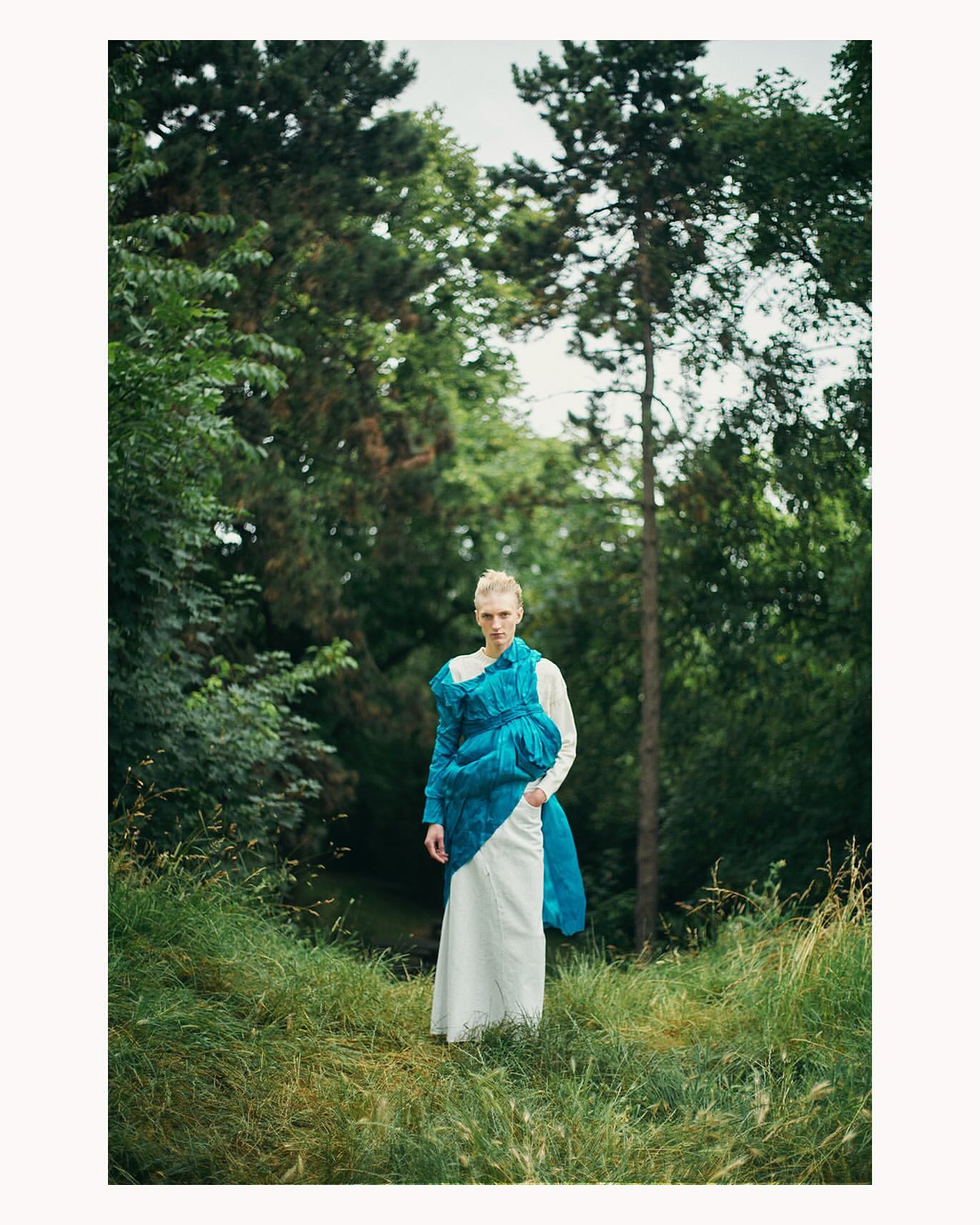 Model standing on a grassy hill wearing a white dress draped in vibrant blue fabric by photographer Ola Rindal.
