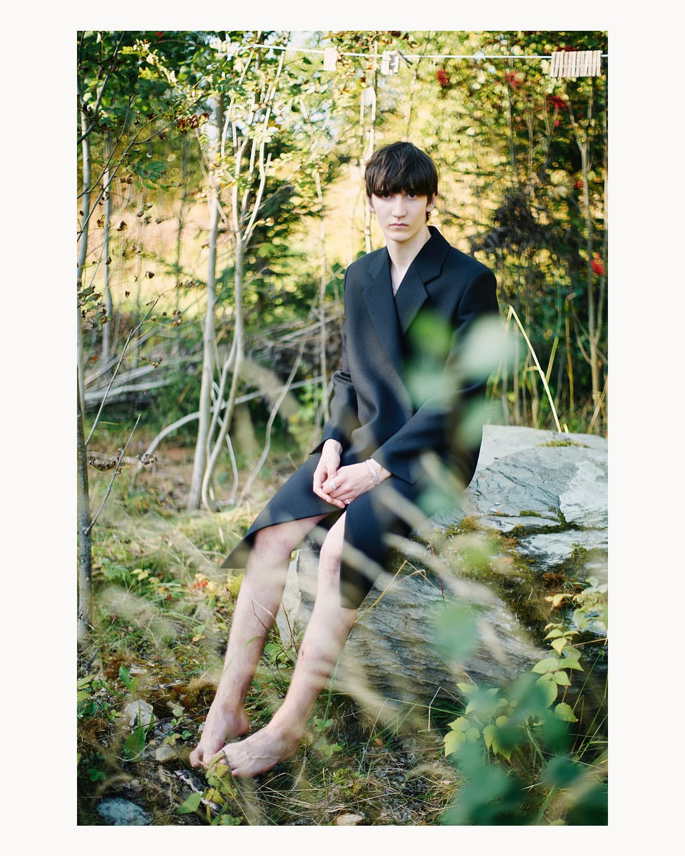 A young adult poses in a black suit in a forest environment, sitting on a rock with bare feet, captured in a natural, editorial style.