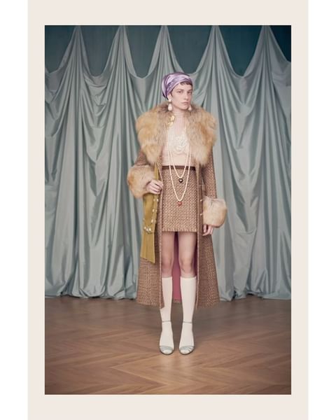 Woman in 1960s-inspired fashion with a fur-collared coat, mini skirt, knee-high boots, and headscarf against a curtain backdrop.
