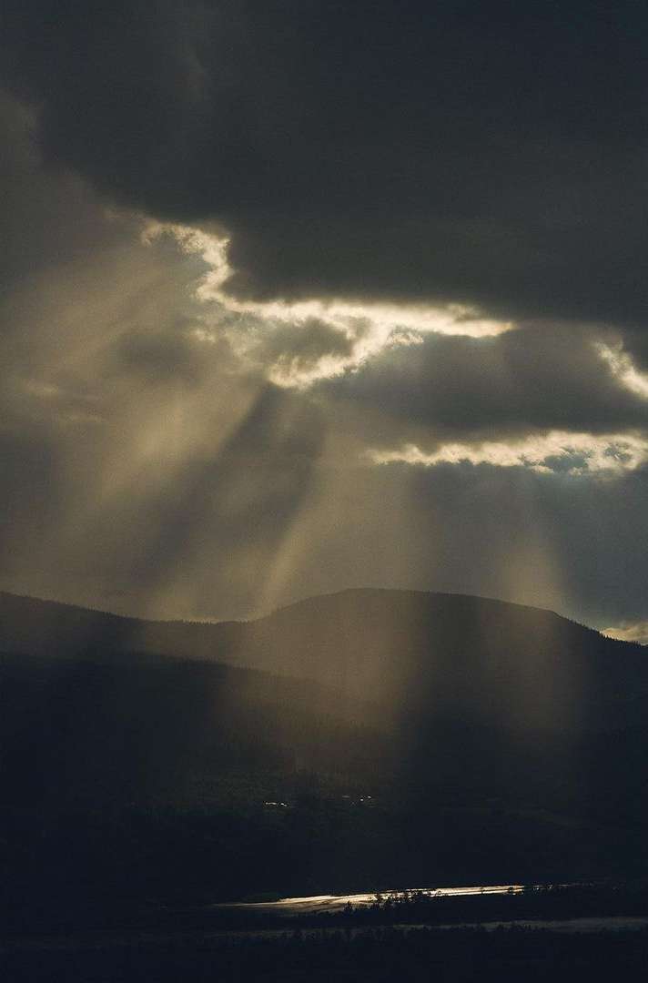 Dramatic mountain landscape with a lake reflecting sunlit rays breaking through dark storm clouds over the scene.