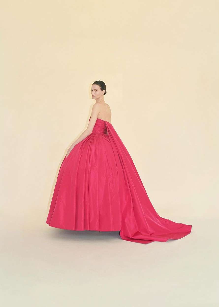 A woman in an elegant fuchsia gown poses in a studio with a light cream backdrop. The dress features a long train and strapless bodice.
