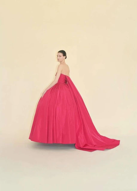 A woman in an elegant fuchsia gown poses in a studio with a light cream backdrop. The dress features a long train and strapless bodice.