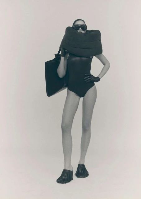 Monochrome fashion shot of a woman in a black bodysuit, oversized eyewear, and fringed shoes holding a large, dark tote bag.