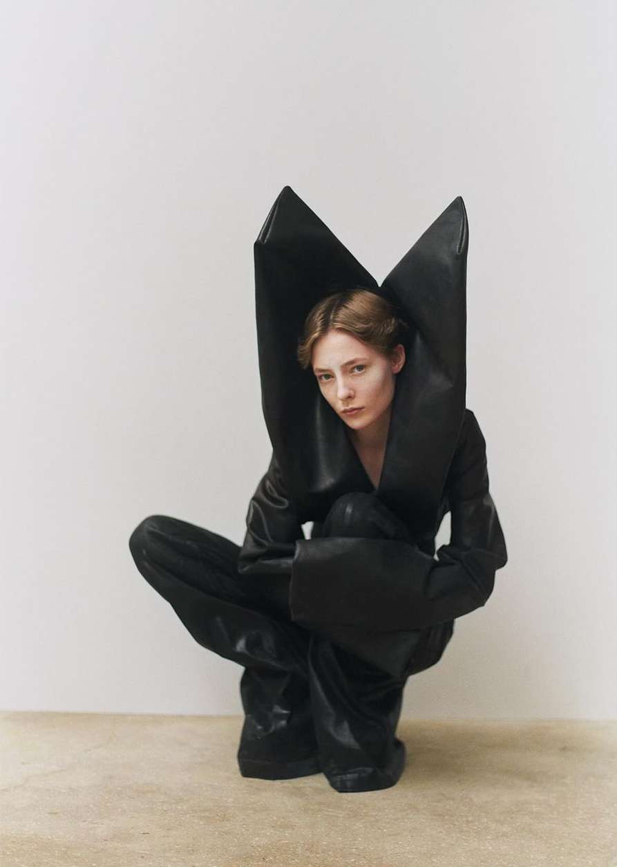 A fair-skinned woman poses in a squat wearing a glossy black outfit, large puffy hood, and matching gloves against a neutral studio backdrop.