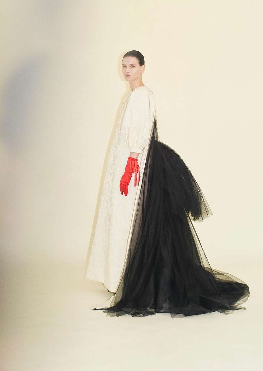 A woman poses in a white dress with a black tulle train and red gloves against a neutral backdrop in a fashion editorial setting.