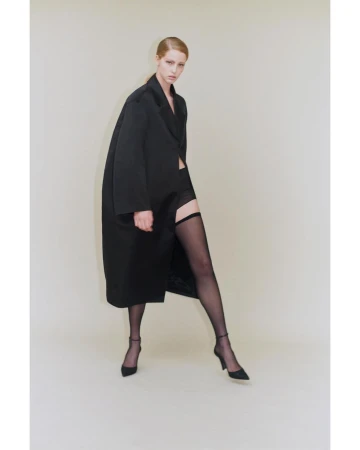 Model in a long black coat wearing sheer stockings and black heels against a neutral studio background by Ola Rindal