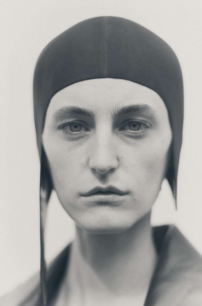 Monochromatic portrait of a person in a close-fitting head covering, gazing directly at the viewer in a muted, neutral-toned setting.