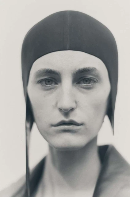 Monochromatic portrait of a person in a close-fitting head covering, gazing directly at the viewer in a muted, neutral-toned setting.