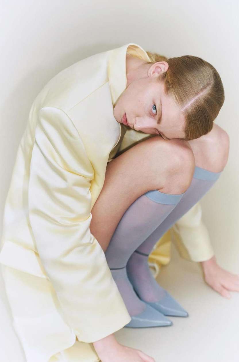 An adult woman in a crouched pose wearing a light yellow jacket, hosiery, and matching shoes in a bright studio setting.